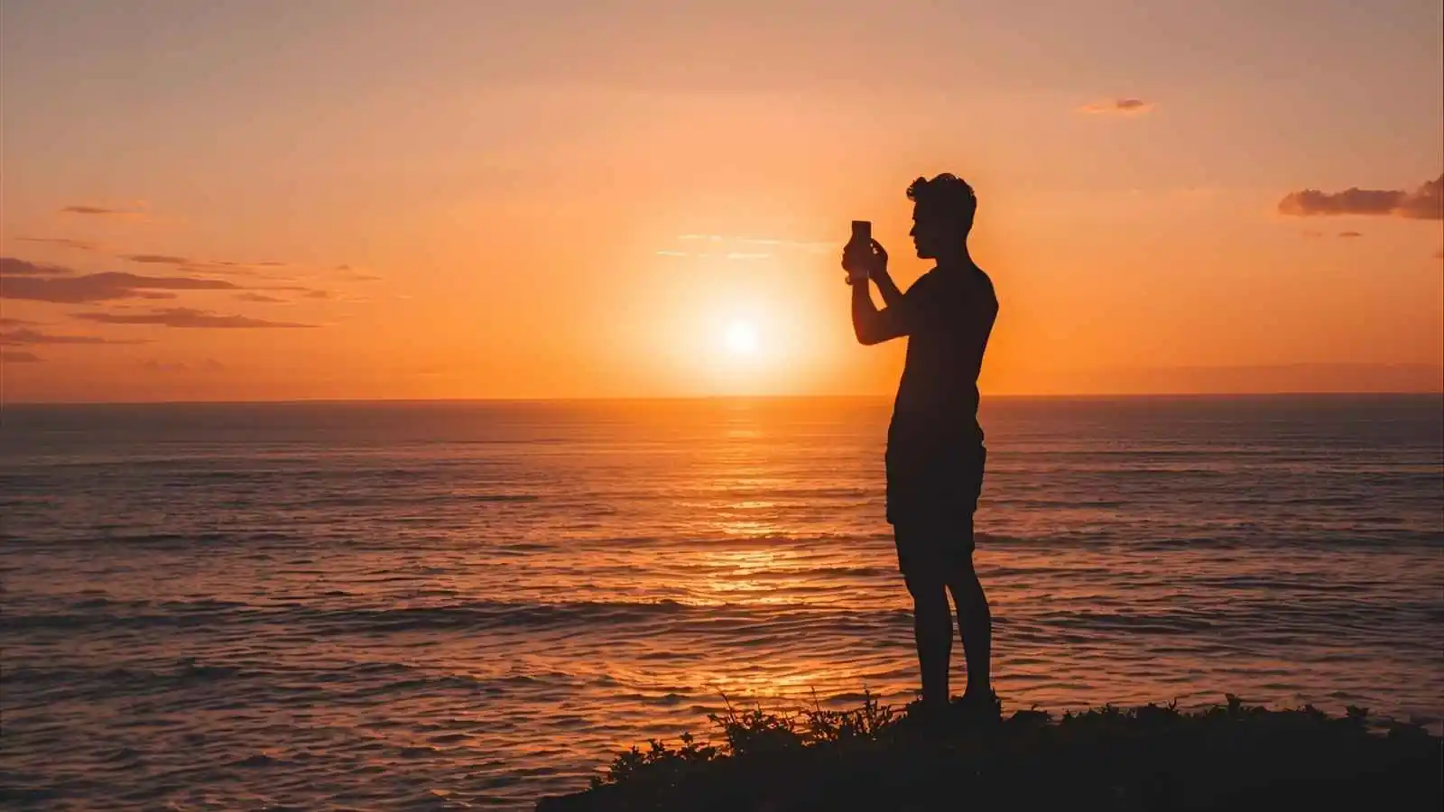 Person taking a photo of a golden sunset over the ocean from a cliff.